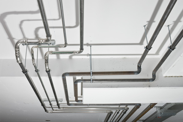 Alt-Text: „Sorgfältig installierte Metallrohre und -verbindungen für ein effizientes Heizungs- und Klimasystem an einer weißen Decke in einem Gewerbebau. - © Viega