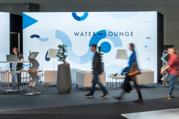 Alt-Text: „Fachbesucher passieren den Messestand der Water Lounge, ausgestattet mit modernen Sitzgelegenheiten und grünen Pflanzen, während einer Veranstaltung zur Präsentation neuester Innovationen in der Sanitär- und Heiztechnik. - © Constantin Meyer / VDS