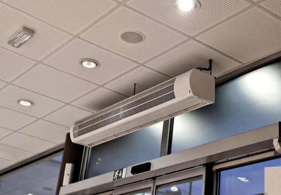 „Luftschleieranlage zur Klimaregulierung oberhalb von Glastüren in einem Gewerbegebäude, umgeben von strukturierten Deckenplatten und integrierter Beleuchtung. - © Bild: Michal - stock.adobe.com