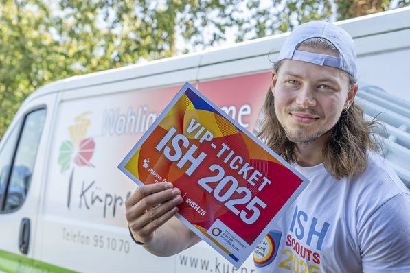 „Ein SHK-Auszubildender lächelt und präsentiert ein ‚VIP Ticket ISH 2025‘ vor einem Firmenfahrzeug. Er trägt eine Schirmmütze und ein T-Shirt mit dem Logo seines Ausbildungsbetriebs im Bereich Sanitär-, Heizungs- und Klimatechnik. - © ZVSHK