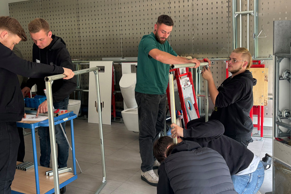 Fünf Personen montieren Metallrahmen in einer Werkstatt. Im Hintergrund sind Werkzeuge, Toiletten und Geräte zu sehen. - © Tece