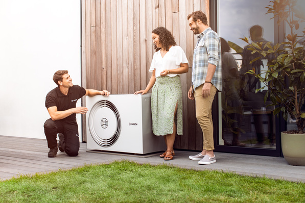 Ein Techniker erklärt einem lächelnden Paar vor ihrem modernen Haus eine Bosch-Wärmepumpe. - © Bosch
