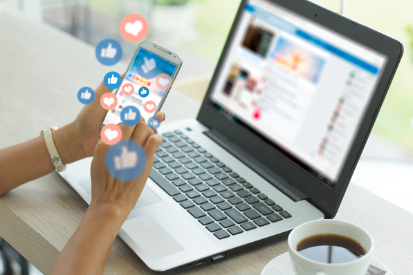Person, die ein Smartphone mit Symbolen für soziale Medien benutzt, neben einem Laptop und einer Tasse Kaffee auf einem Schreibtisch. - © Urupong - stock.adobe.com